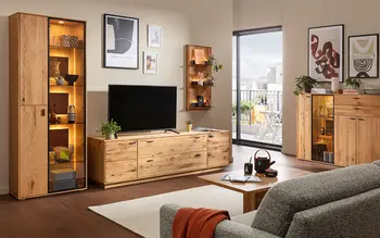 Gemütliches Wohnzimmer mit modernen Holzmöbeln, beleuchteter Vitrine, TV-Lowboard, Wandregal, Sideboard und grauem Sofa, Blick auf Balkon im Hintergrund.